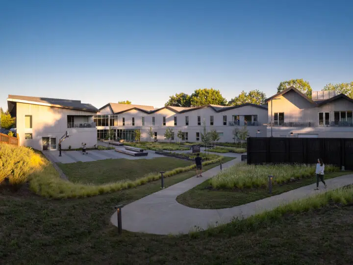 A view of Fora Health from the back part of the lush, green courtyard, looking down at a winding path that leads to the active area of the courtyard, which features a basketball court, various seating options, and a garden.