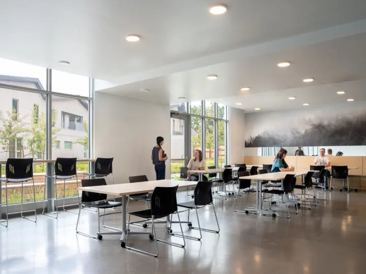 Several people sit at rolling tables in a bright, daylit cafeteria.