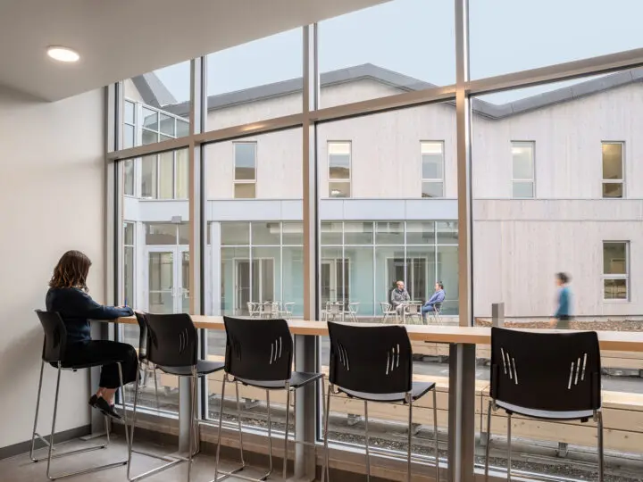 A person sits on a tall chair at a window-facing bar, looking onto the outdoor patio.