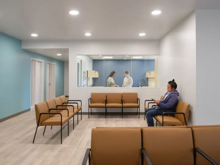 A patient sits in a small reception area with rust-orange chairs and mellow blue walls.