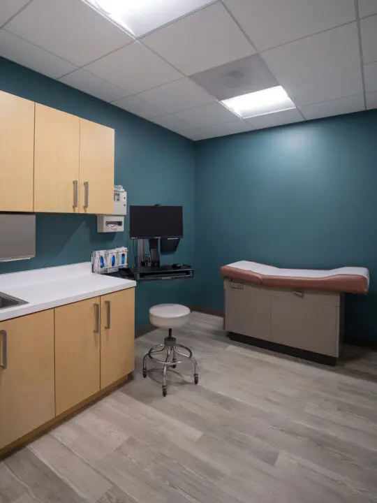 A small consultation room is equipped with an exam table, a wall-mounted workstation, a rolling stool, medical supplies, and wood cabinets.