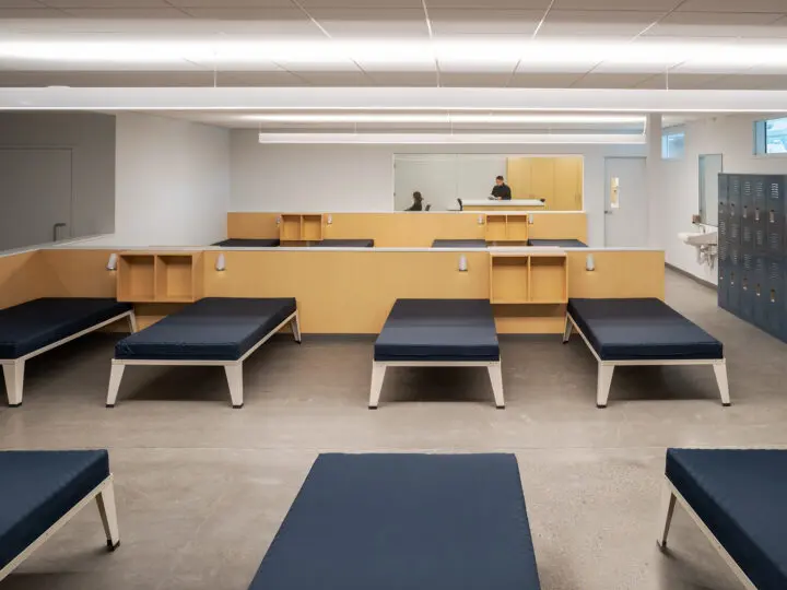A white-walled room of beds is partitioned by low wood walls with built-in storage and lighting.