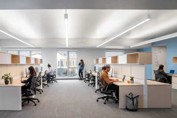 Several rows of wooden desks with beige dividers and overhead storage are being utilized by administrative staff.