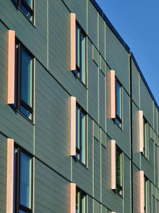 A close-up view of the louvered residential windows showcases the vertical copper fins that protrude from the horizontal sage-green siding at each opening.