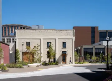 A view of the exterior of the renovated the space between PRAx, the heating plant, and McAlexander Fieldhouse, shows a planted landscape in front of the restored historic Rehearsal Classroom Building, which features white-painted brick and wooden doors.