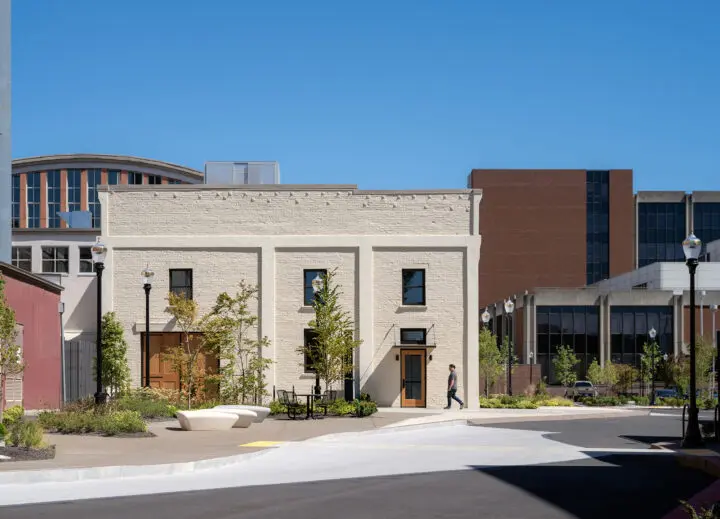 A view of the exterior of the renovated the space between PRAx, the heating plant, and McAlexander Fieldhouse, shows a planted landscape in front of the restored historic Rehearsal Classroom Building, which features white-painted brick and wooden doors.