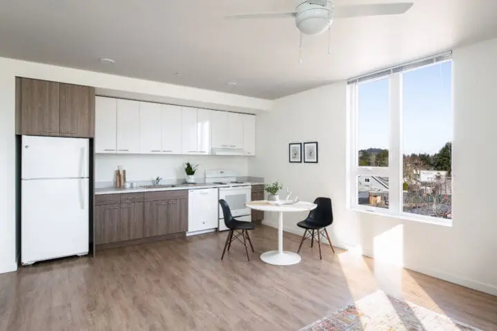 A light-filled kitchen in one of 72Foster’s units features a small dining table, brown lower cabinets, white upper cabinets, and white appliances.
