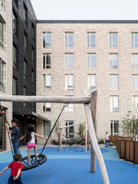 Three kids play outside on a wooden swing structure in a fenced-in outdoor play area at The Aurora.