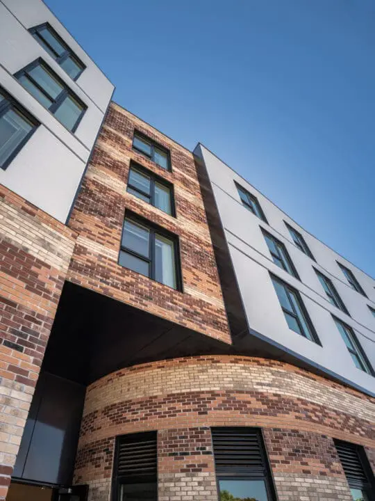 Speckled bands of earth-toned brickwork and tilted planes of white siding meet at various angles of an apartment building.