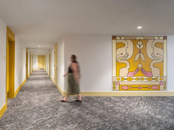A woman in a long skirt is captured walking through a white-walled and gray-carpeted hallway lined with bright yellow apartment doors.