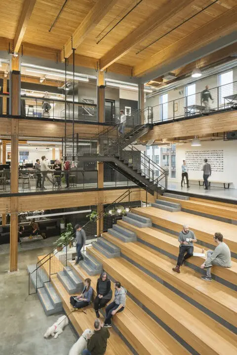 Looking down from above, people converse on the large wooden bleacher stairs in the atrium.