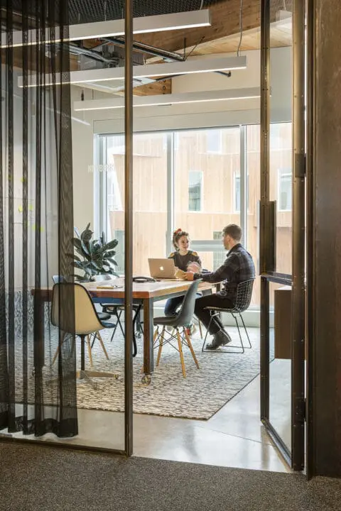 Two people converse in a glass-walled meeting room over a wooden worktable and a diverse selection of chairs.