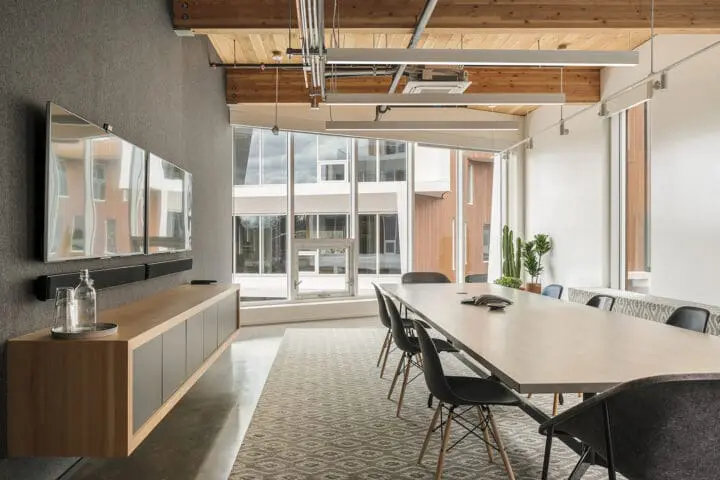 Looking through a linear meeting space, a long wooden table with black chairs above a grey, ikat-patterned carpet below a floor-to-ceiling aperture of windows.